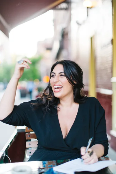 Roey laughing as she writes in a notebook at a sidewalk table.
