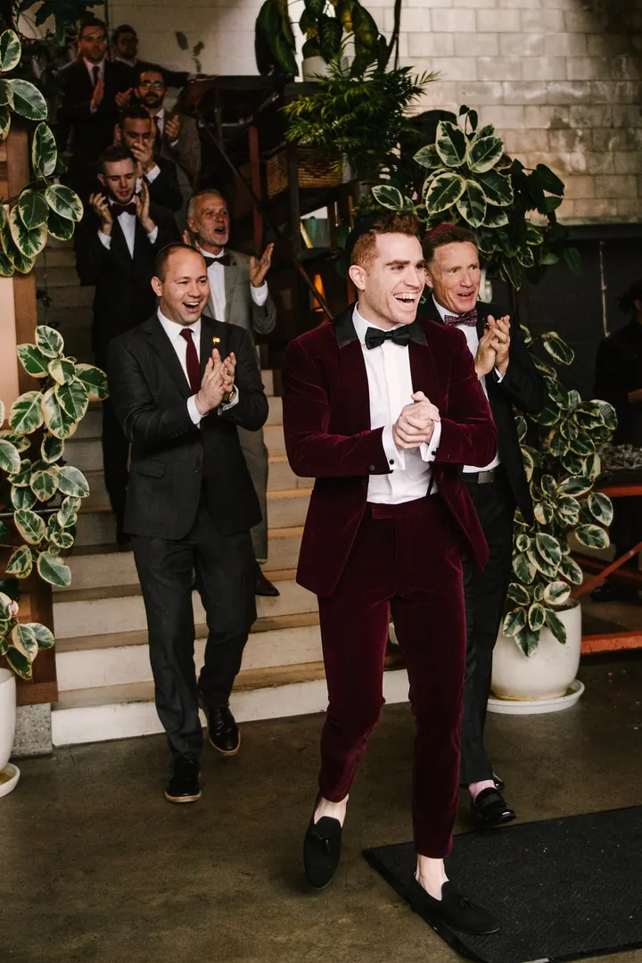 A groom in a burgundy velvet tuxedo jacket smiles as guests applaud behind him on a staircase.
