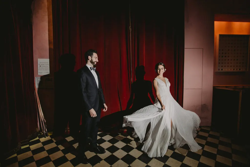 A bride in a pleated gown twirls in front of a red velvet curtain on a checkerboard floor while the groom watches.