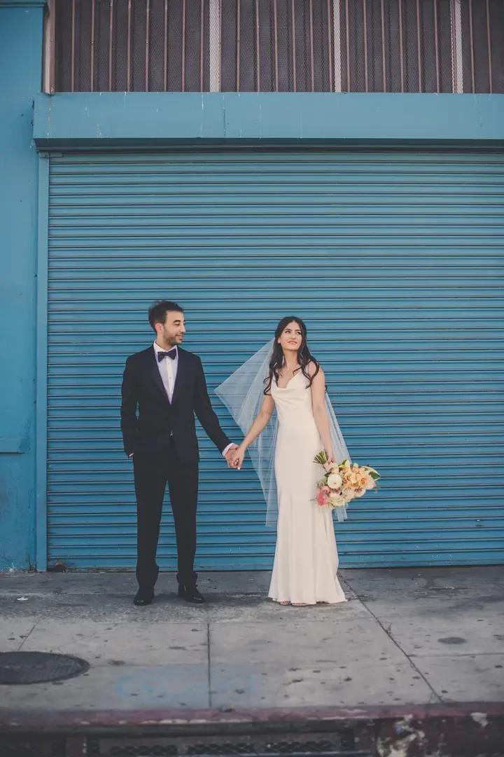 A bride and groom hold hands in front of a teal roll-up door, her veil lifting in the breeze.